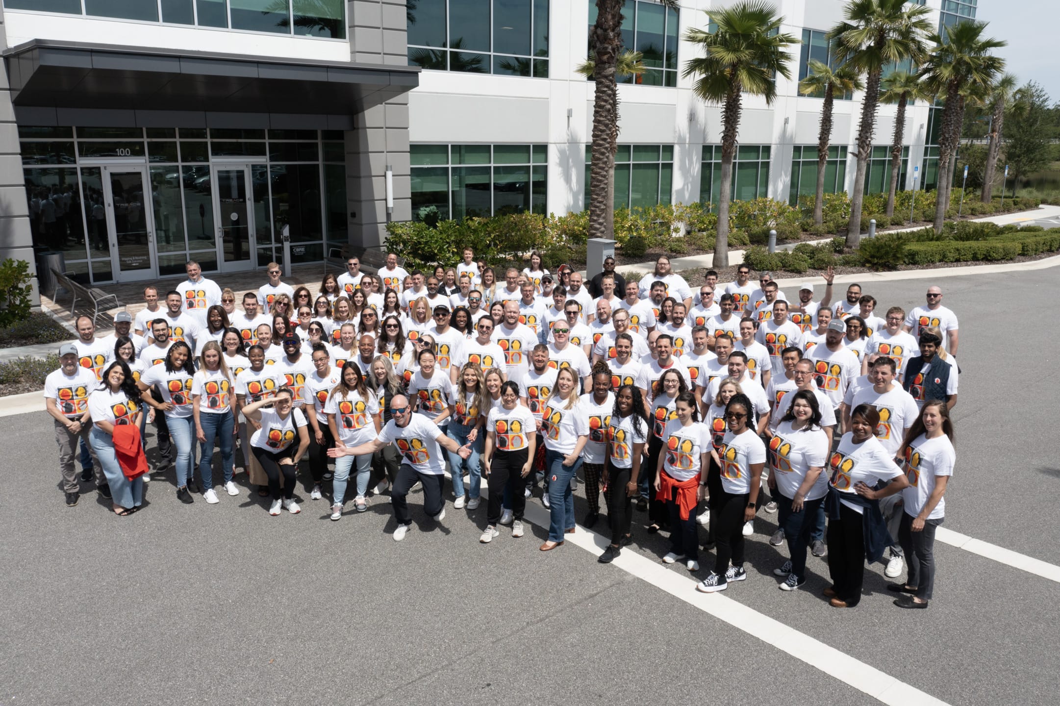 Group of people wearing matching white T-shirts with colorful designs, posing in front of a modern building with palm trees.