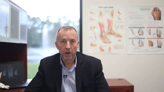 A medical professional in a blazer and button-down shirt speaking in an office, with Lapiplasty bunion correction anatomical posters and a laptop showing X-rays in the background.