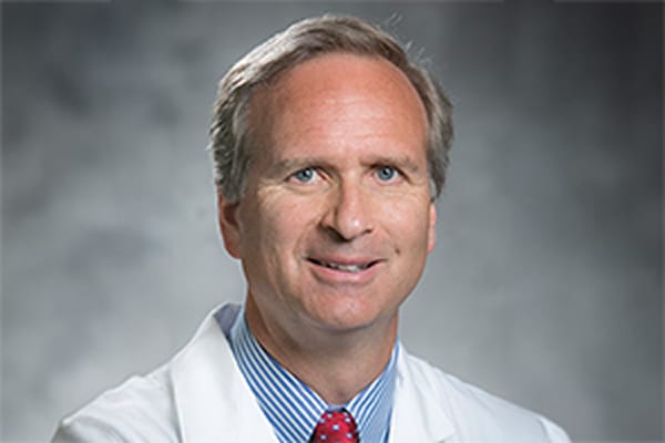 Smiling man in a white lab coat and striped shirt against a gray backdrop.