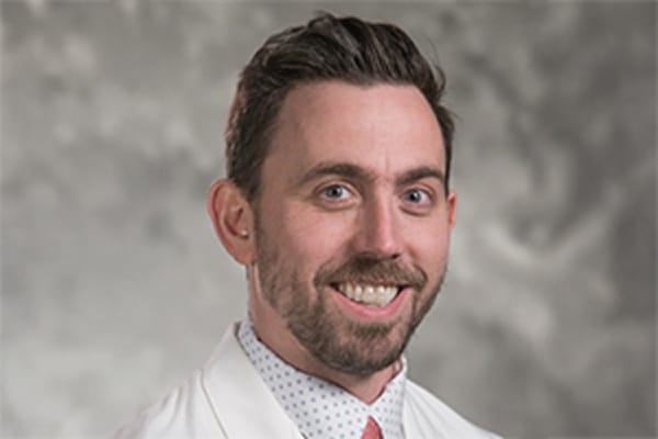 Smiling person with short hair wearing a white coat against a gray background.