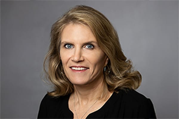 Professional headshot of a woman with blonde hair wearing a black top and smiling, set against a gray background.
