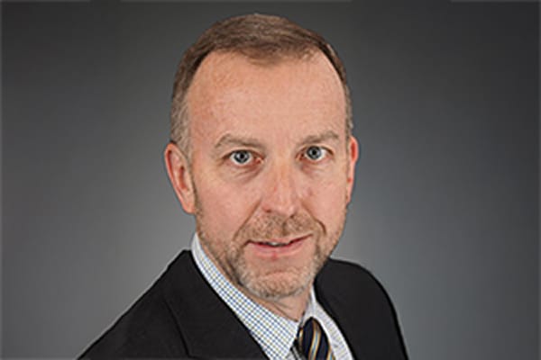 Man in a suit with a gray background, appearing professional and confident, looking directly at the camera.
