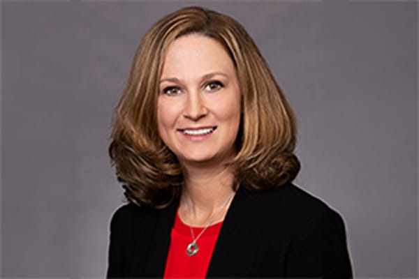 Person with shoulder-length brown hair in a black blazer and red shirt, smiling against a gray background.