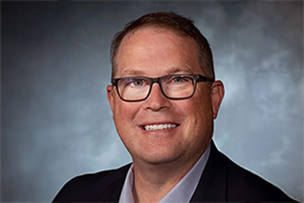 Smiling person wearing glasses and a dark jacket against a cloudy blue background.