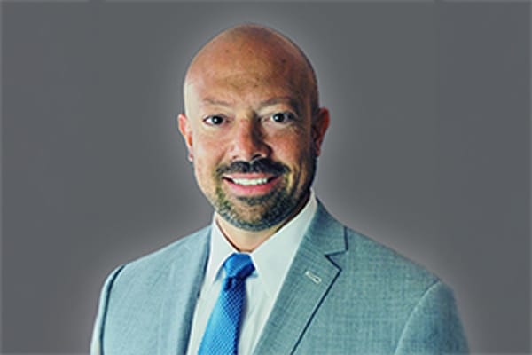 Man in a light gray suit and blue tie smiling against a gray background