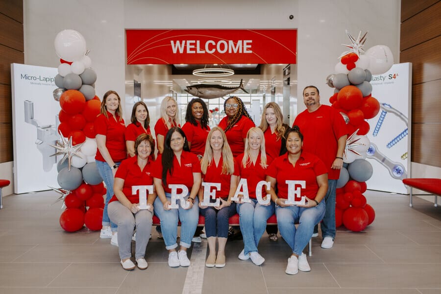 Team of people in matching red shirts holding letters spelling TREACE, in a modern lobby with balloons and welcome sign.