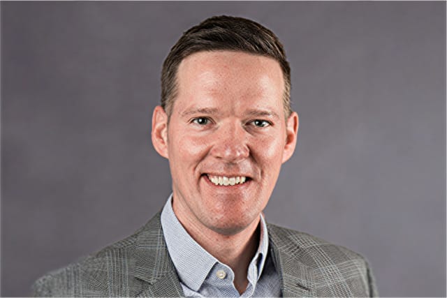 Portrait of a smiling man in a gray blazer and blue shirt against a gray background.