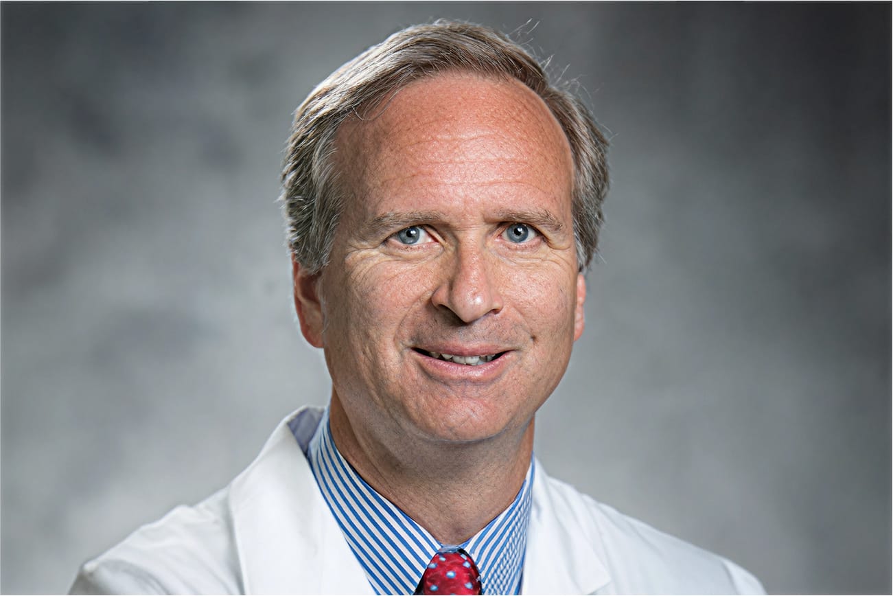 Smiling person in a white coat with a blue striped shirt and red tie against a gray background.