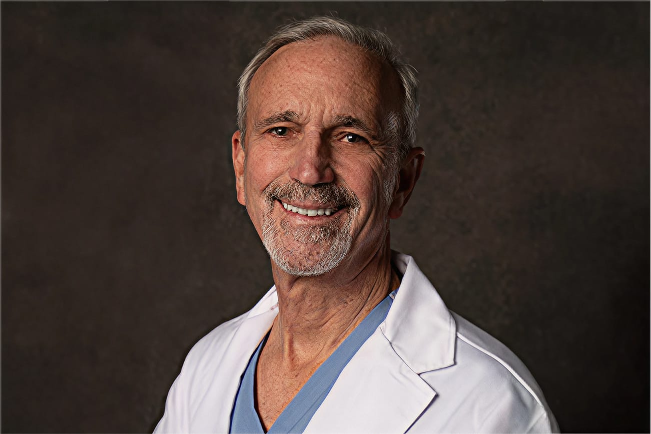 Smiling older person in a white coat and blue shirt against a dark background.