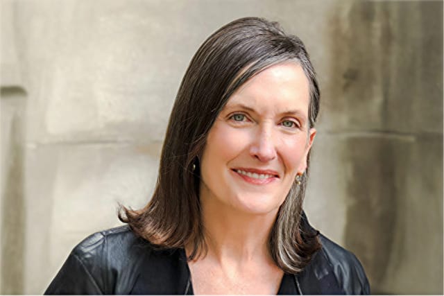 Person with gray hair and a black jacket smiling against a neutral background.