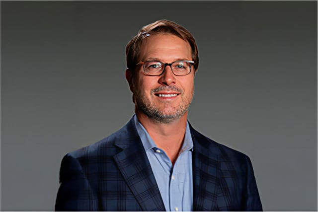 Man wearing glasses and a blue checkered suit jacket smiling against a gray background.