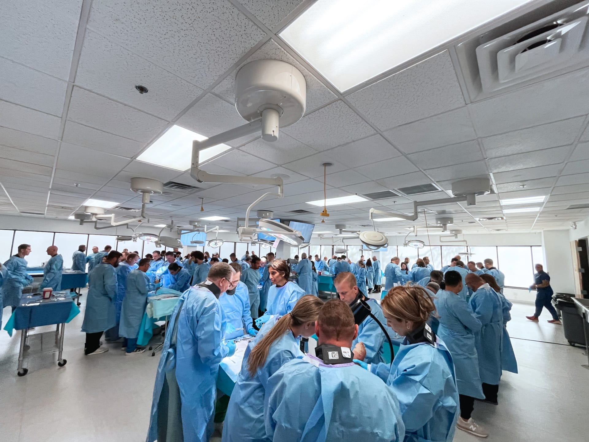 Medical professionals in blue scrubs collaborate in a busy hospital room filled with equipment and overhead lighting.