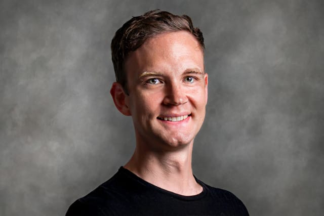 Smiling person with short hair wearing a black shirt against a gray background.