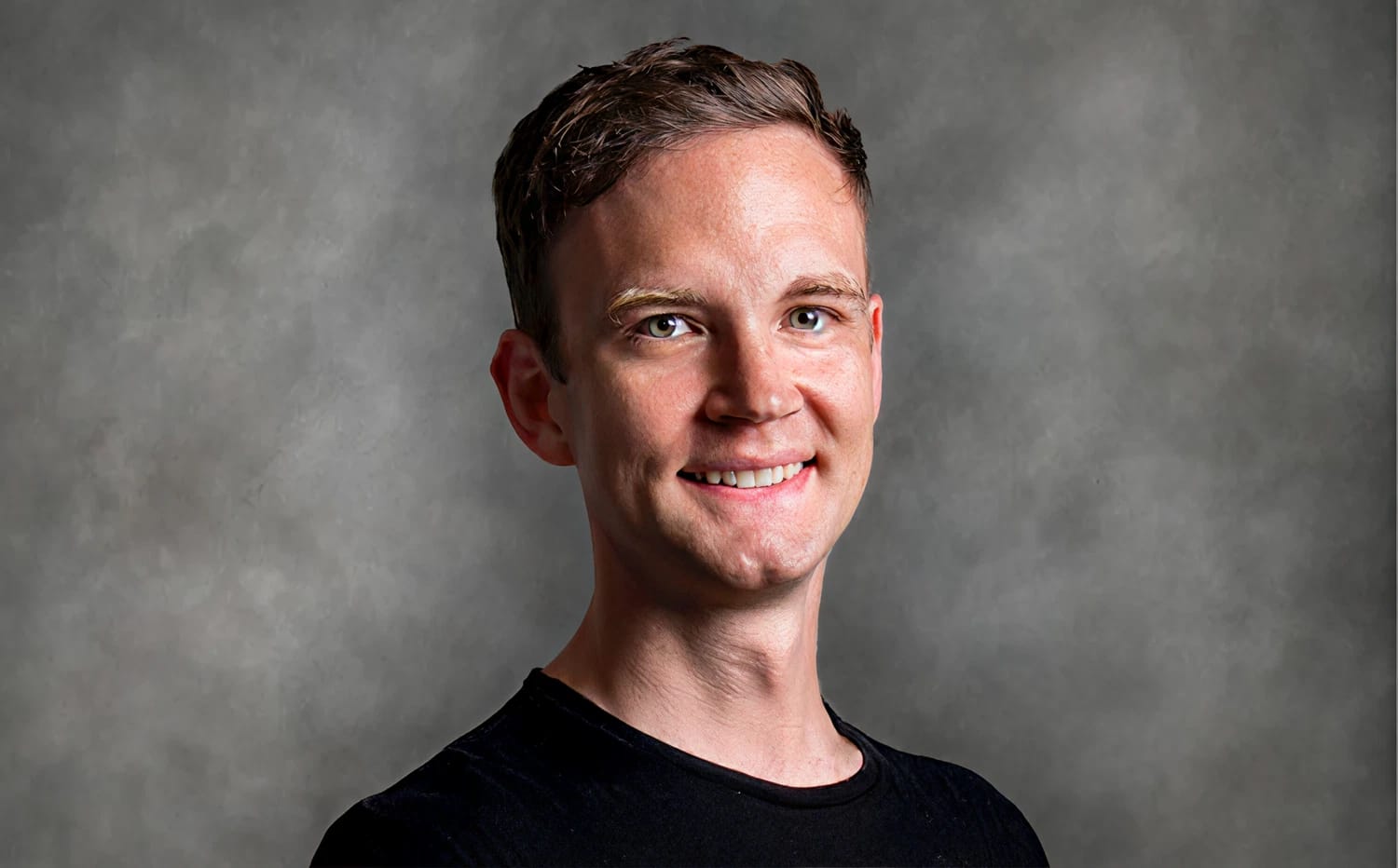 Smiling person with short hair wearing a black shirt against a gray background.
