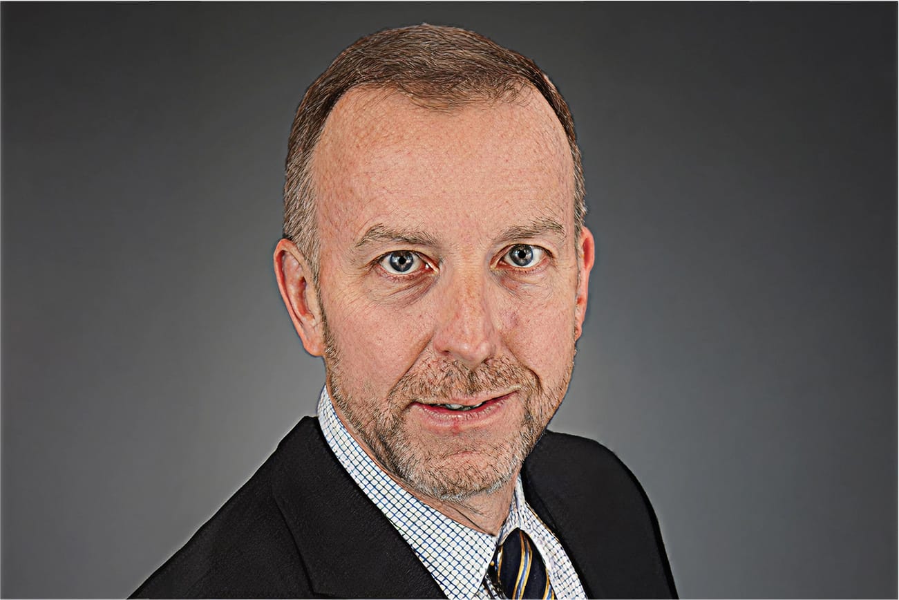 Man in formal attire with a suit and tie, neutral expression, against a gray background