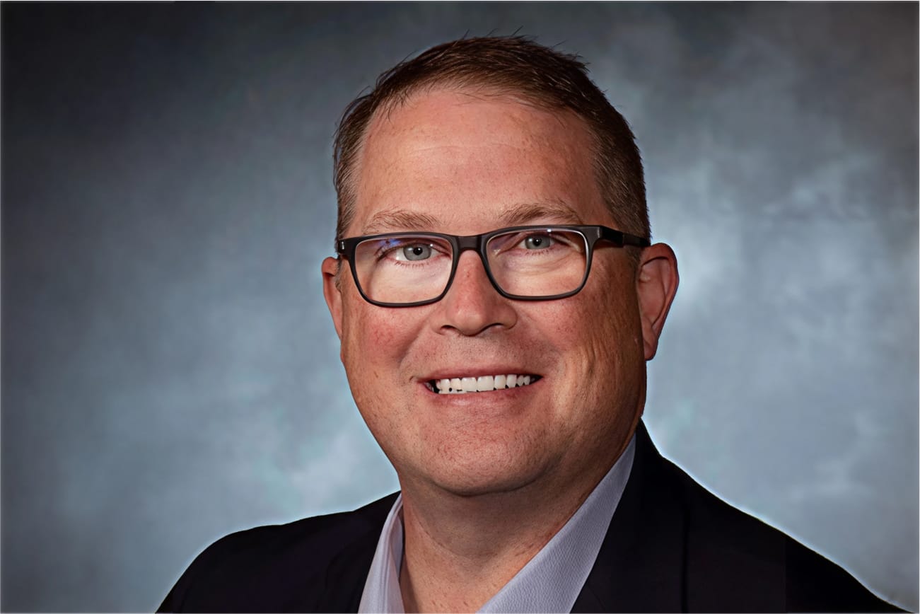 Man with glasses and short hair smiling against a gray background.