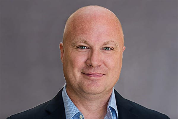 A bald man in a suit and blue shirt smiling against a gray background.