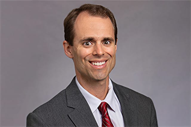 Smiling man in a suit with a red tie, against a gray background.