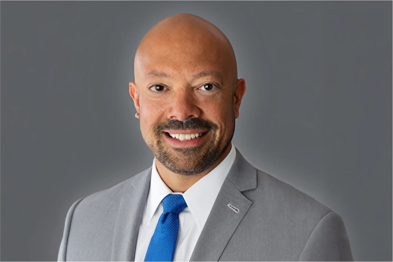 Man in a gray suit and blue tie smiling against a gray background.