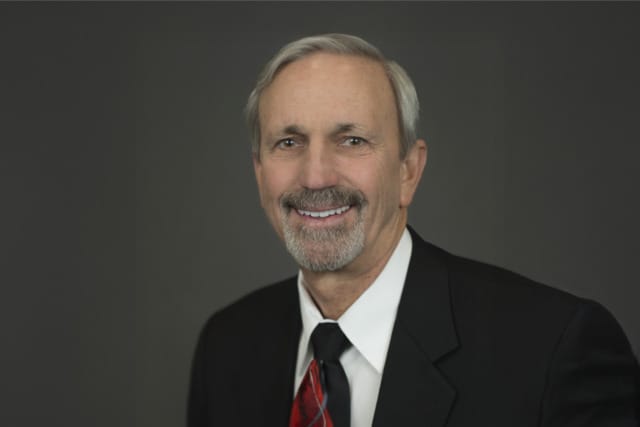 Smiling man in a black suit and red tie against a dark gray background.