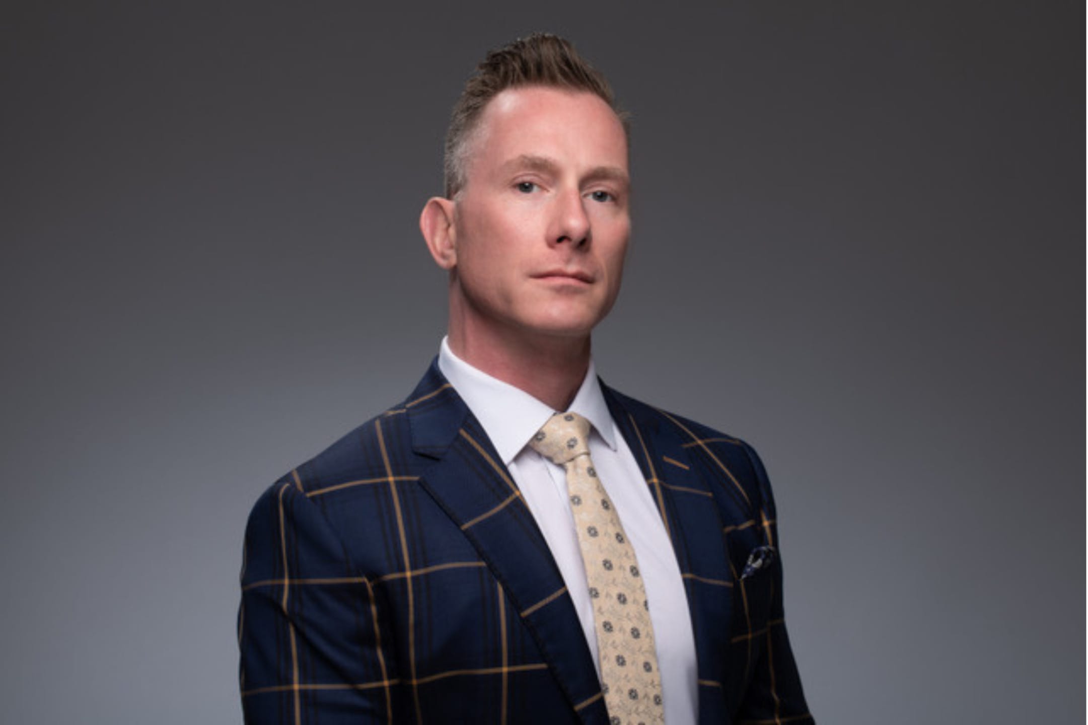 Man wearing a checkered blue suit and beige tie against a gray background.