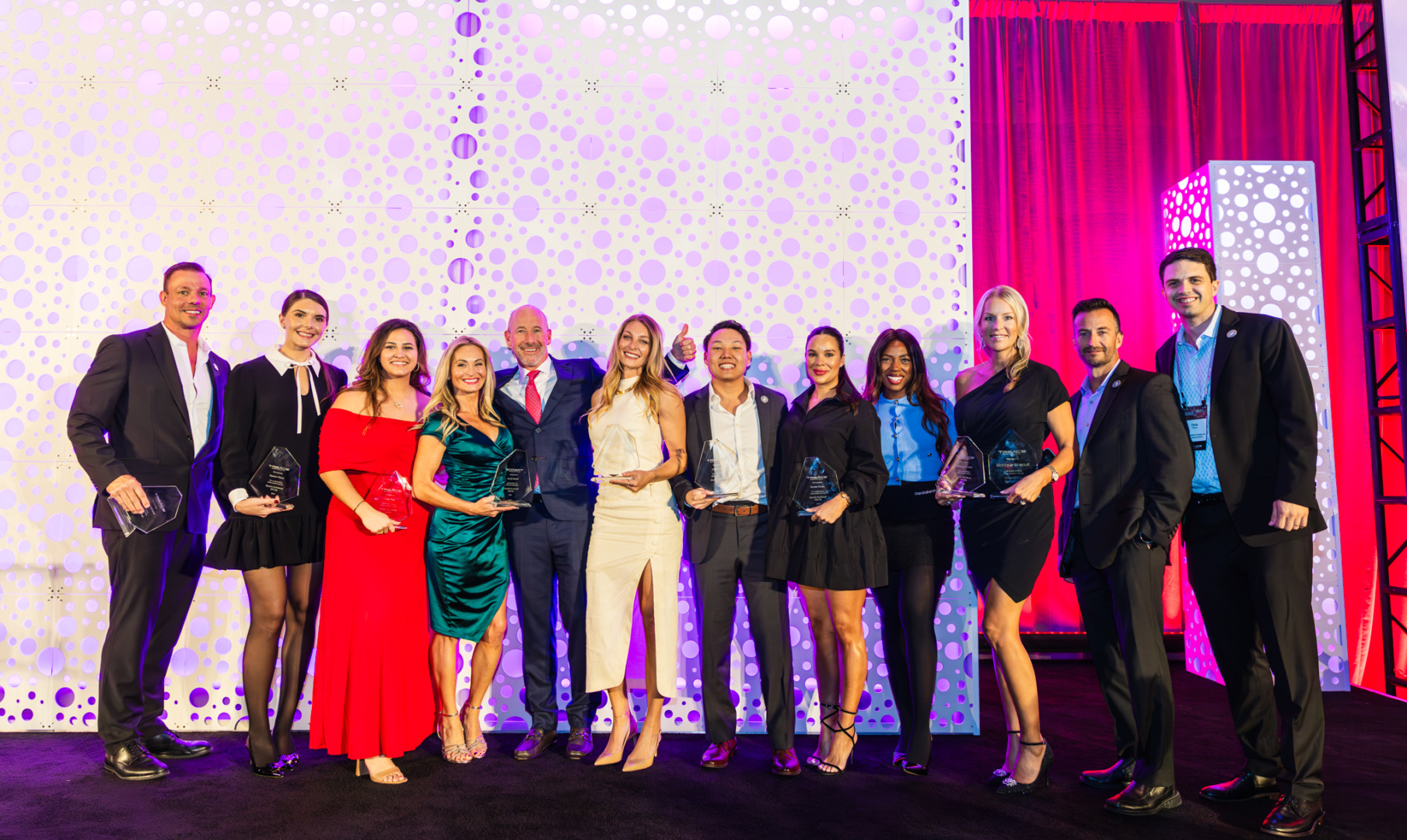Group of people in formal attire holding awards on a stage with decorative lighting.