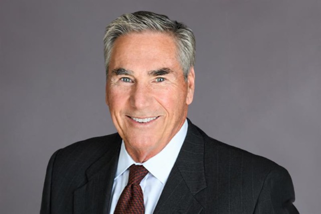 Man in a suit with a gray background, smiling and having salt-and-pepper hair.