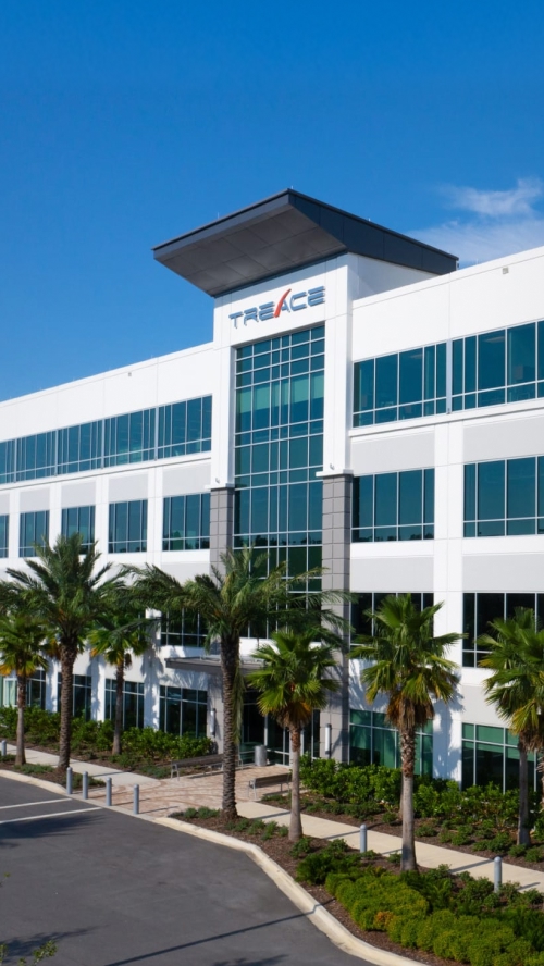 Modern office building with palm trees, large windows, and a sign on the top reading "TREACE," under a clear blue sky.